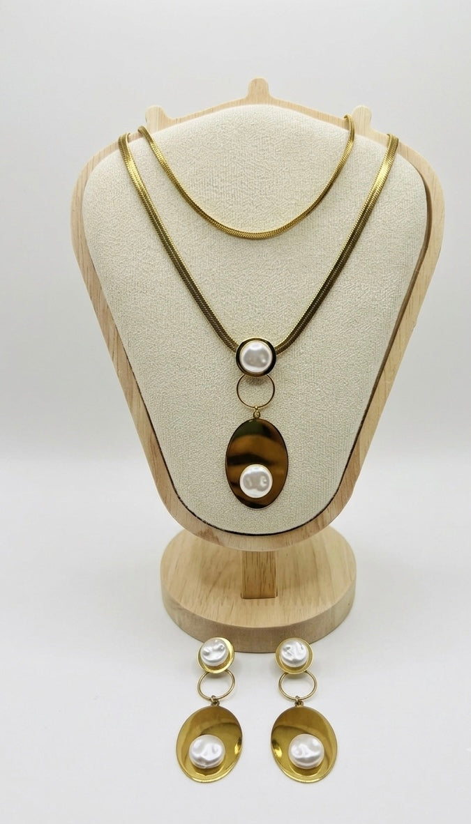 Gold-tone jewelry set featuring a layered necklace and matching drop earrings displayed on a cream jewelry bust stand. The necklace has a thin top chain and a longer pendant with a large oval tiger's eye and two white faux pearls.