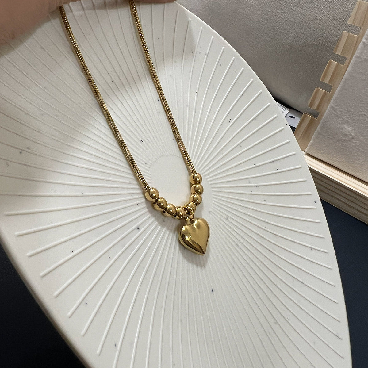 Flat lay view of a gold plated heart pendant necklace featuring spherical beads on a white textured decorative plate