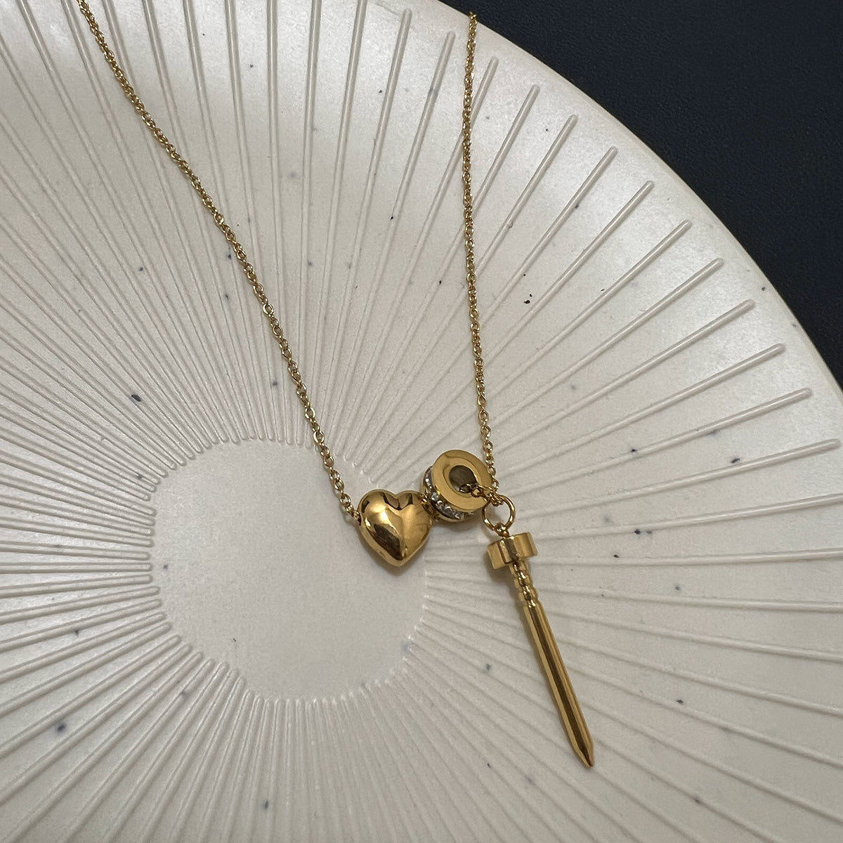 Angled view of a multi-charm anti-tarnish gold necklace with a polished heart and vertical nail pendant on a jewelry stand