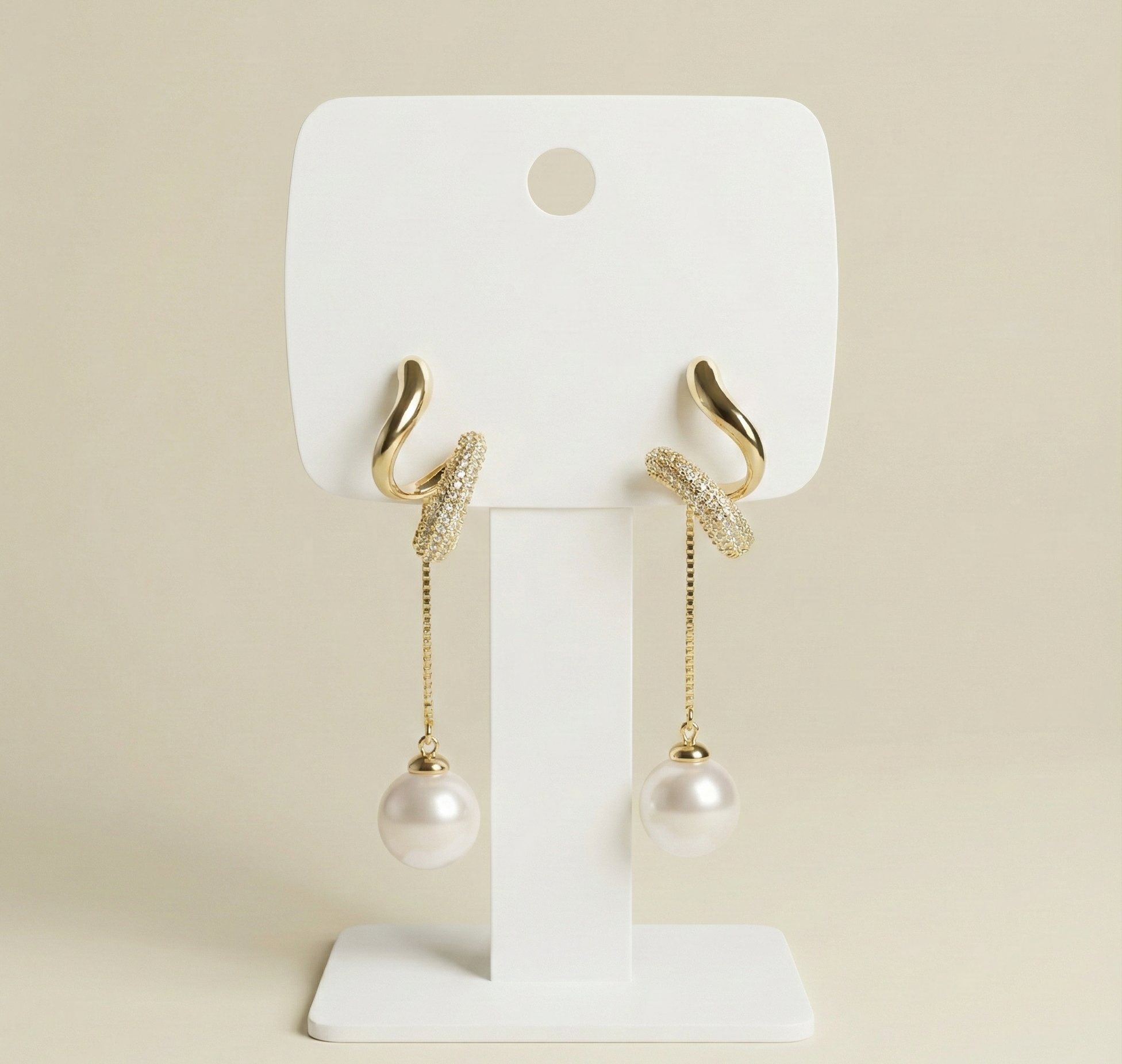 Gold plated curved earrings featuring crystal-studded detailing and a hanging white pearl drop on a white stand