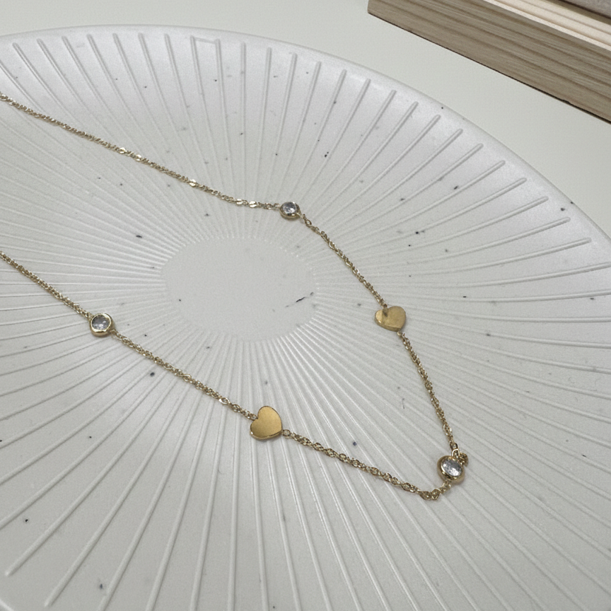 Dainty gold plated station necklace featuring alternating heart charms and clear crystals on a white textured plate
