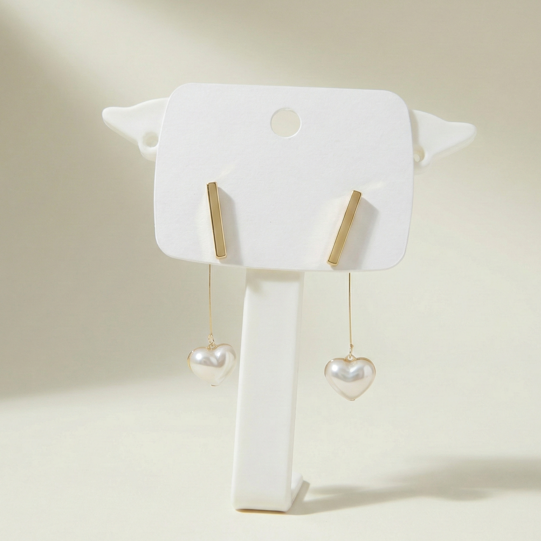 Polished gold bar stud earrings featuring hanging heart-shaped pearls on a minimalist white display stand - zwelzy designs