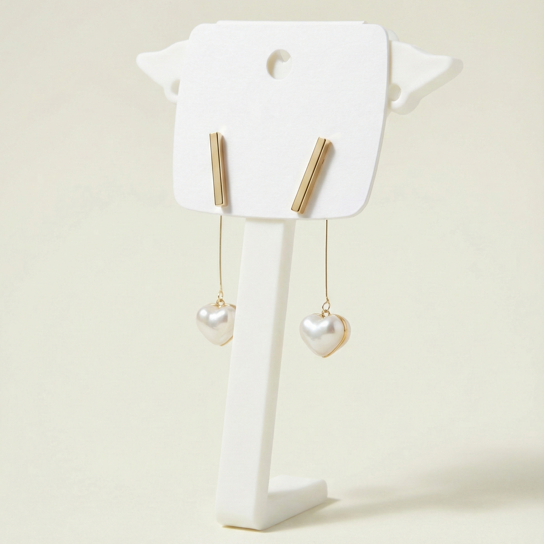 Polished gold bar stud earrings featuring hanging heart-shaped pearls on a minimalist white display stand.