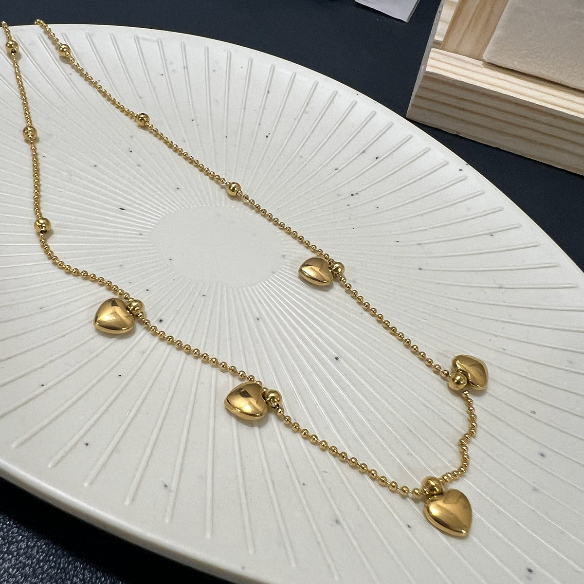 High-shine gold plated ball chain necklace featuring five dangling heart charms on a white textured plate