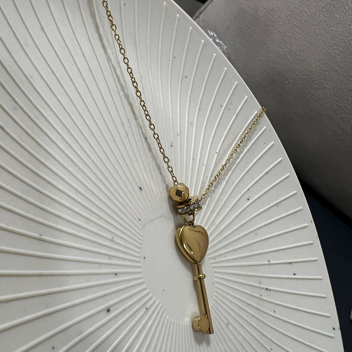 Dainty gold plated heart key pendant necklace resting on a minimalist white sunburst textured plate