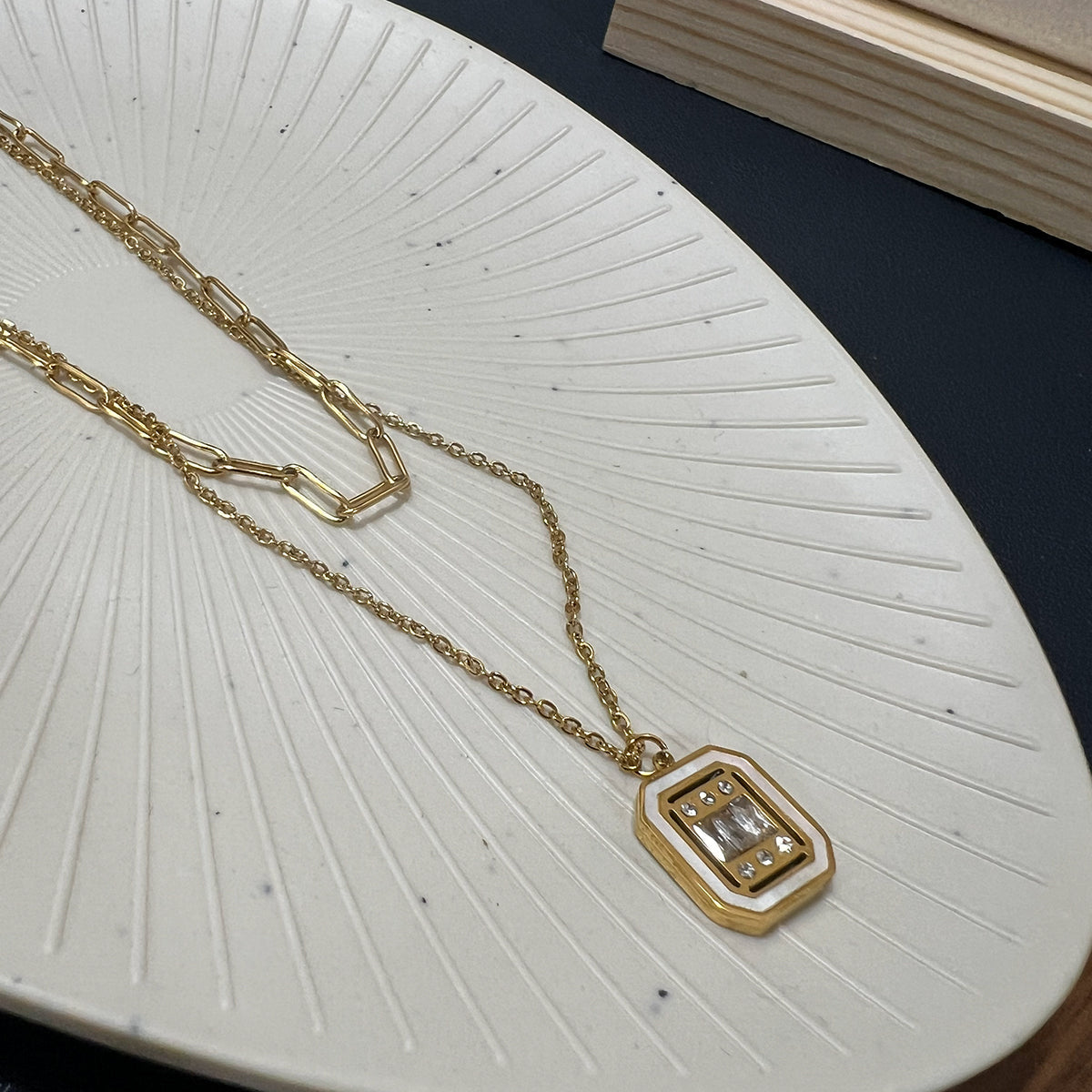 Flat lay of a gold plated layered necklace featuring a paperclip chain and a crystal-set rectangular pendant on a white plate