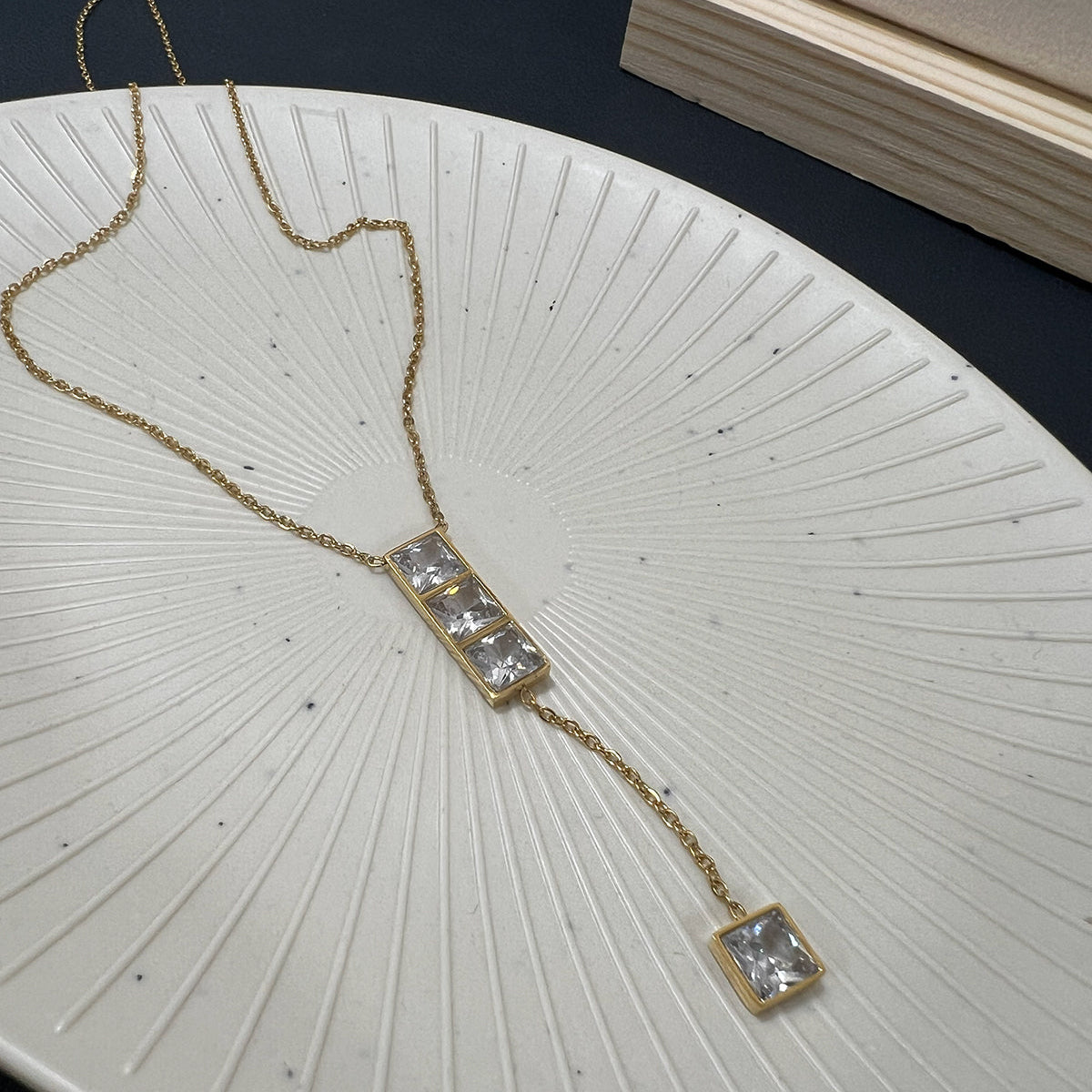 Elegant gold plated lariat necklace featuring three stacked square crystals on a textured white plate
