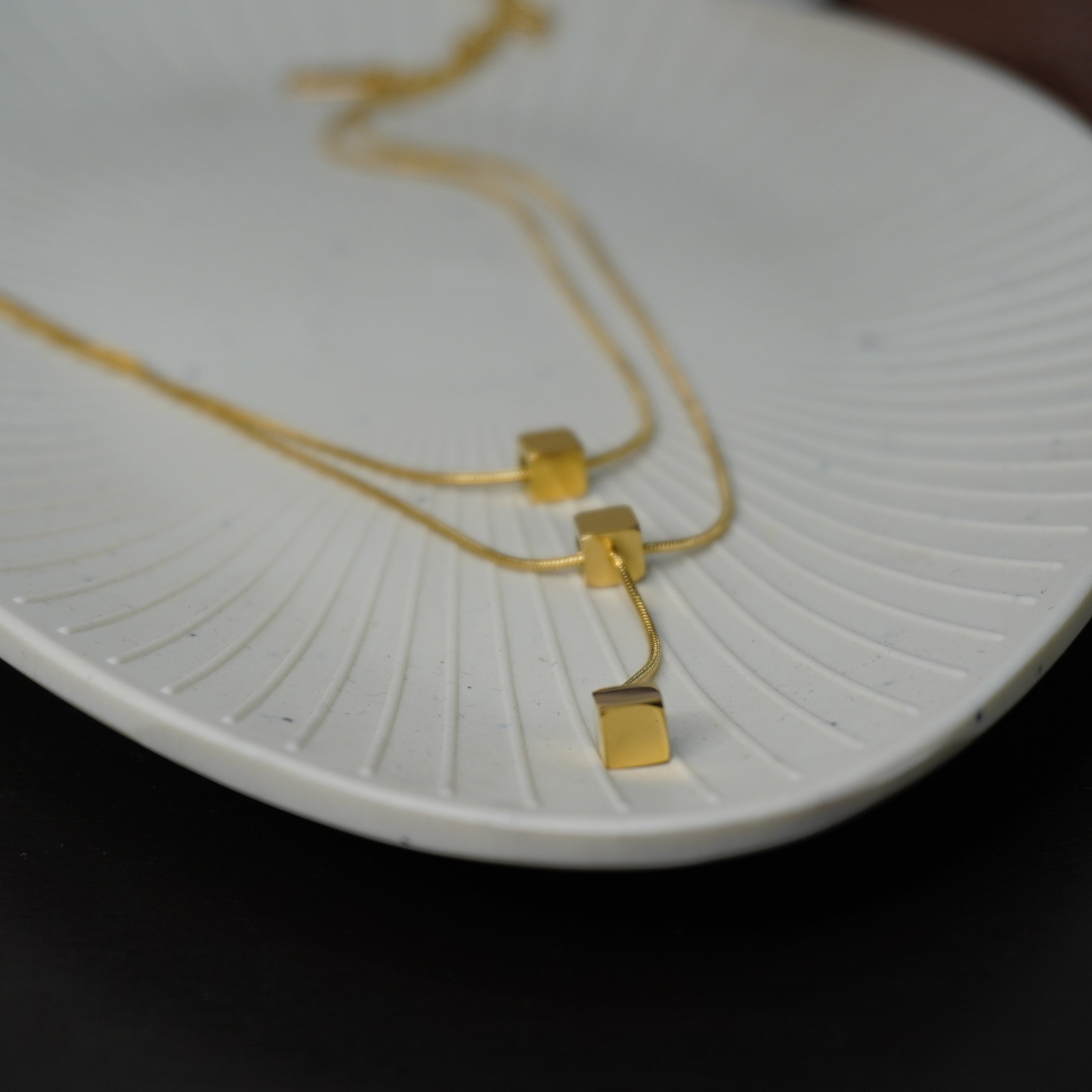 Top view of a dual layer gold plated cube necklace resting on a white textured ceramic plate.