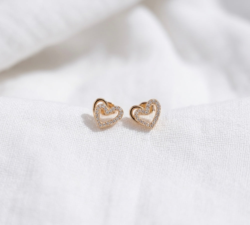 Pair of gold-plated stud earrings in an interlocking heart design, featuring shimmering pavé crystals, resting on a soft white cotton fabric background with bright, natural lighting.