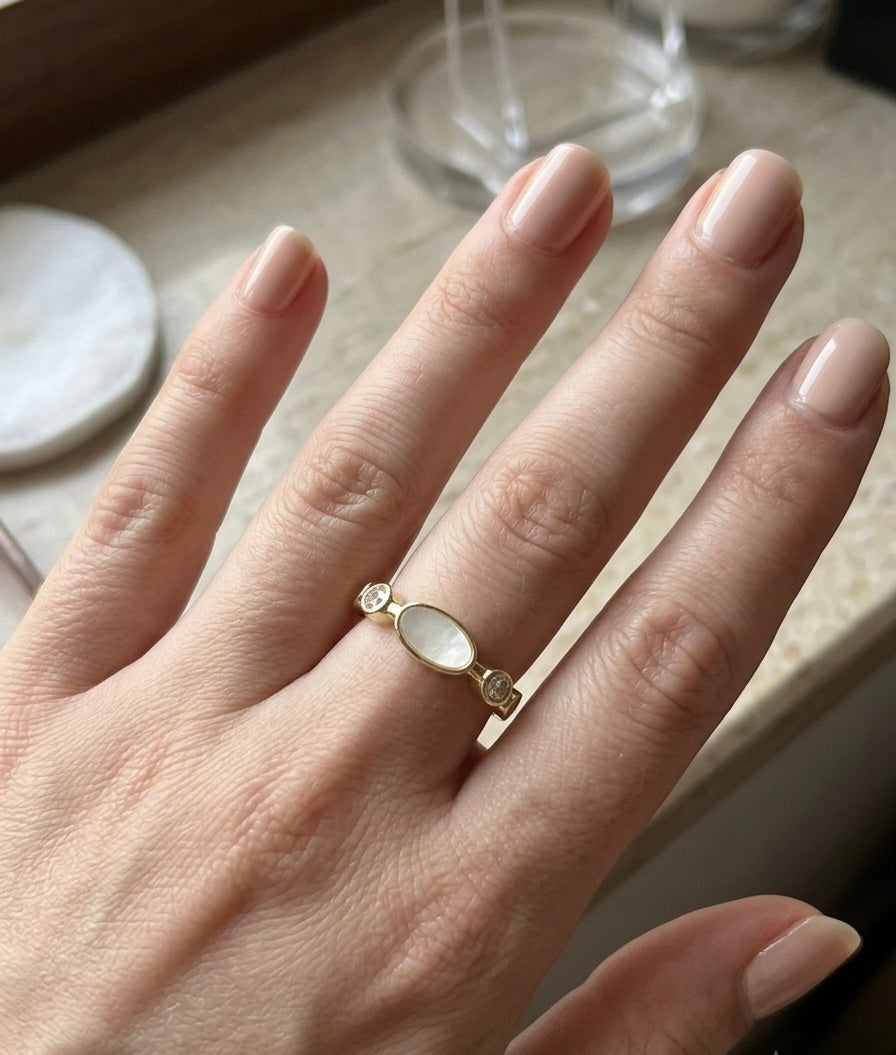 A gold ring with an oval white stone and small clear accent stones, worn on a hand with neutral manicured nails.