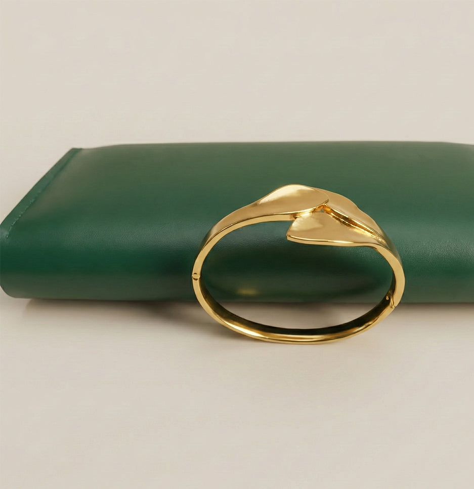 Side view of an elegant gold leaf crossover bangle made of stainless steel placed near a green leather clutch
