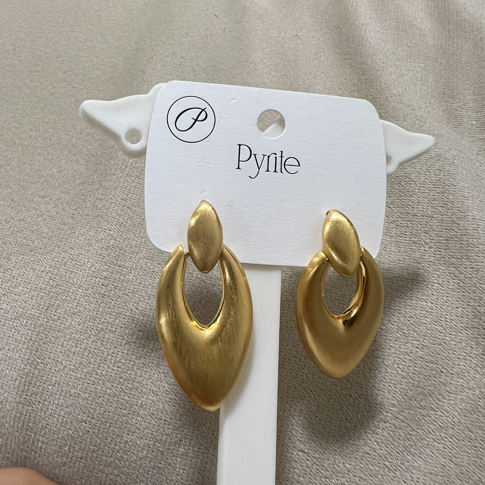 Pair of sculptural brushed gold statement earrings standing on a minimalist white pedestal against a neutral background