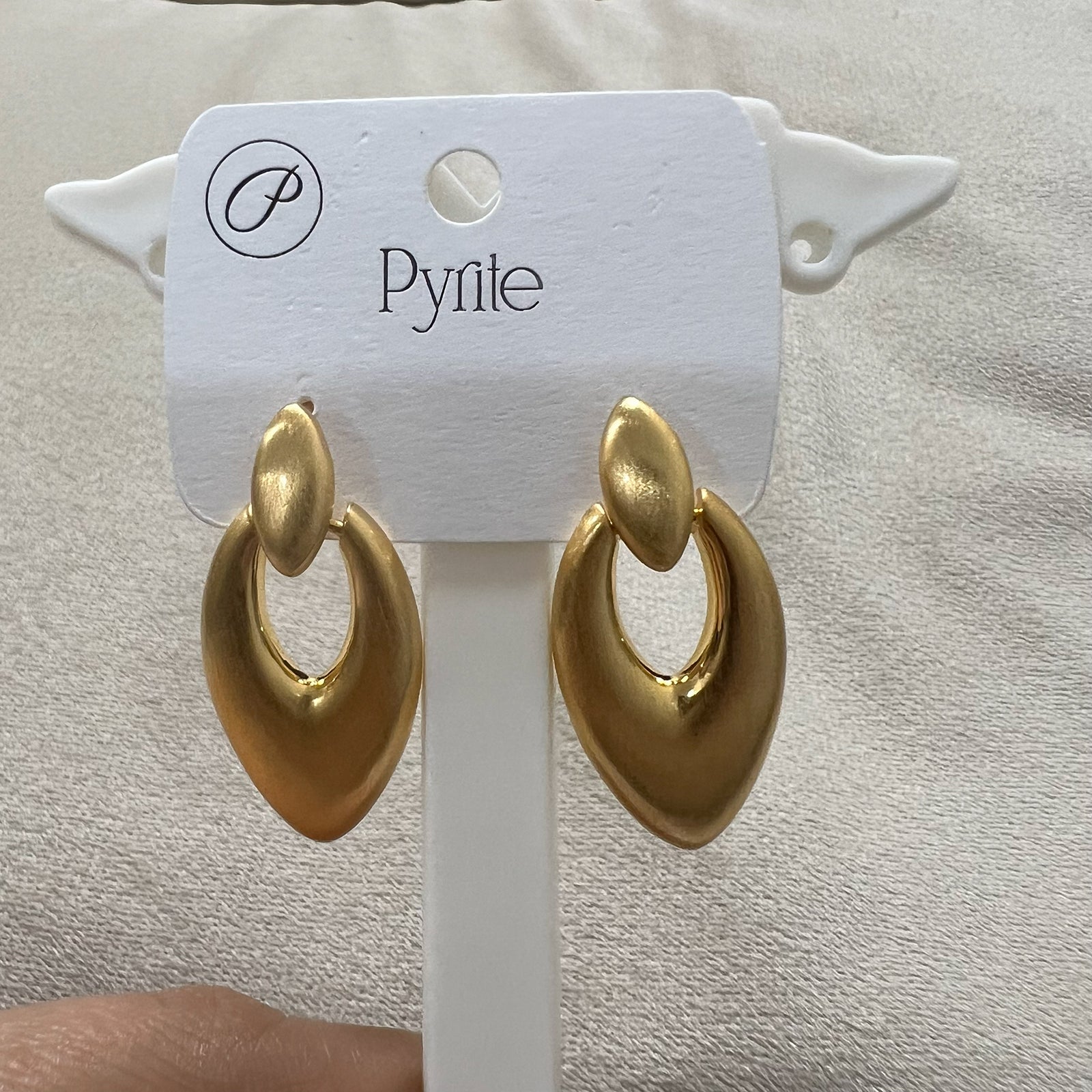 Pair of sculptural brushed gold statement earrings standing on a minimalist white pedestal against a neutral background