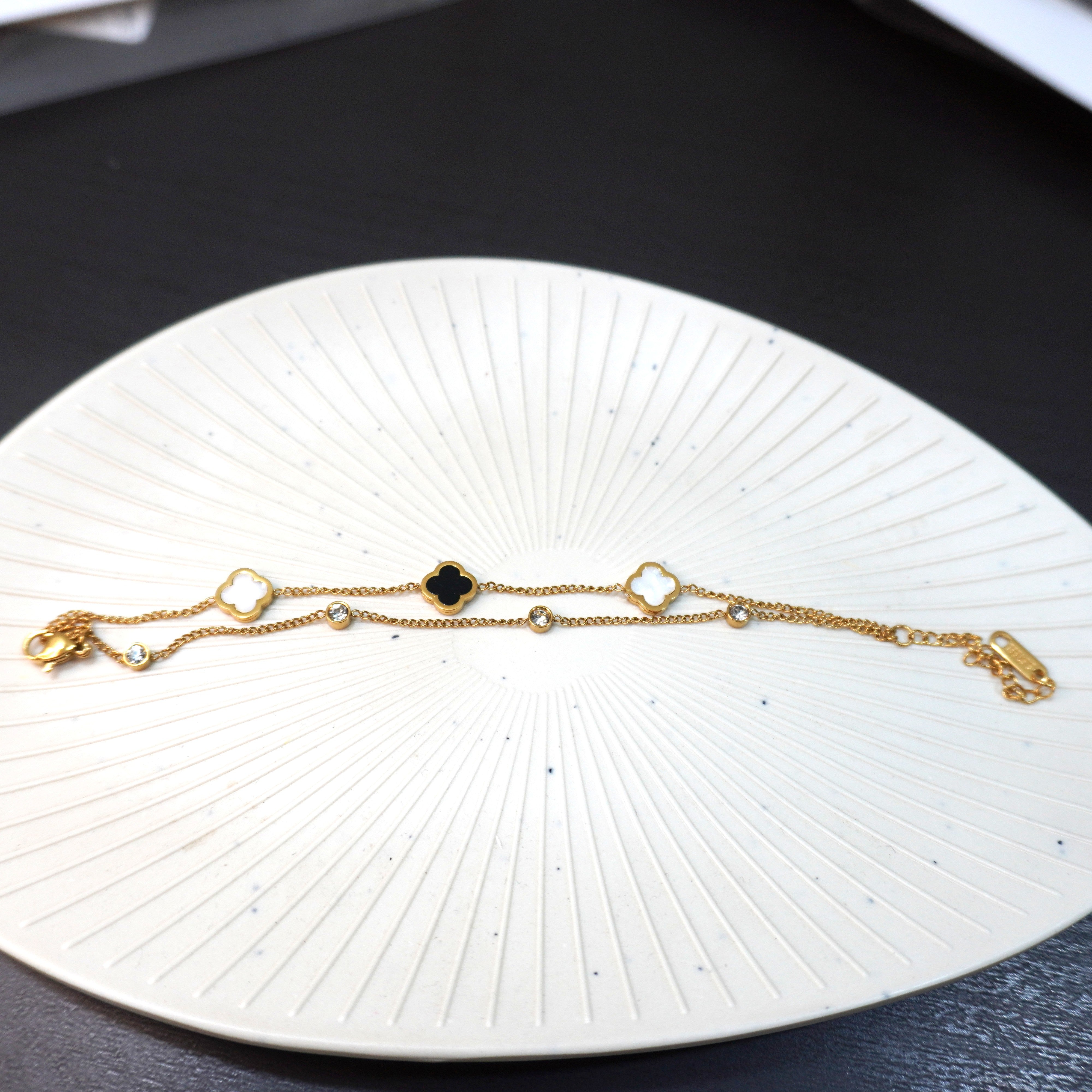Gold plated dual layer bracelet featuring black and white clover charms on a white textured decorative plate.