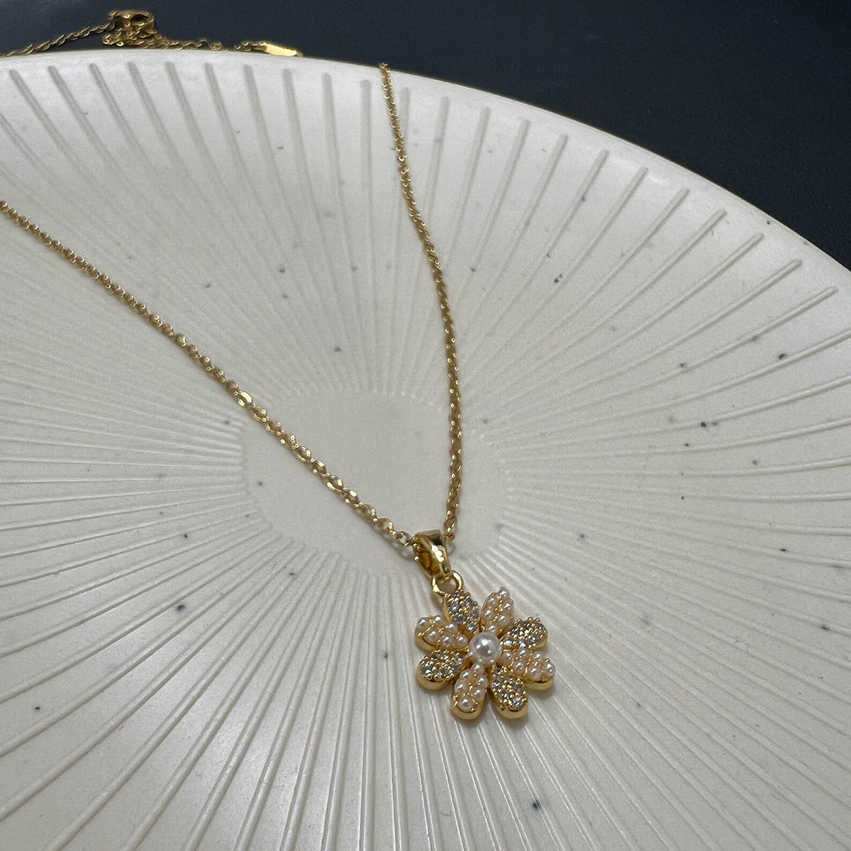 Close-up of a gold plated flower pendant featuring pearls and crystals placed on a white sunburst textured plate.