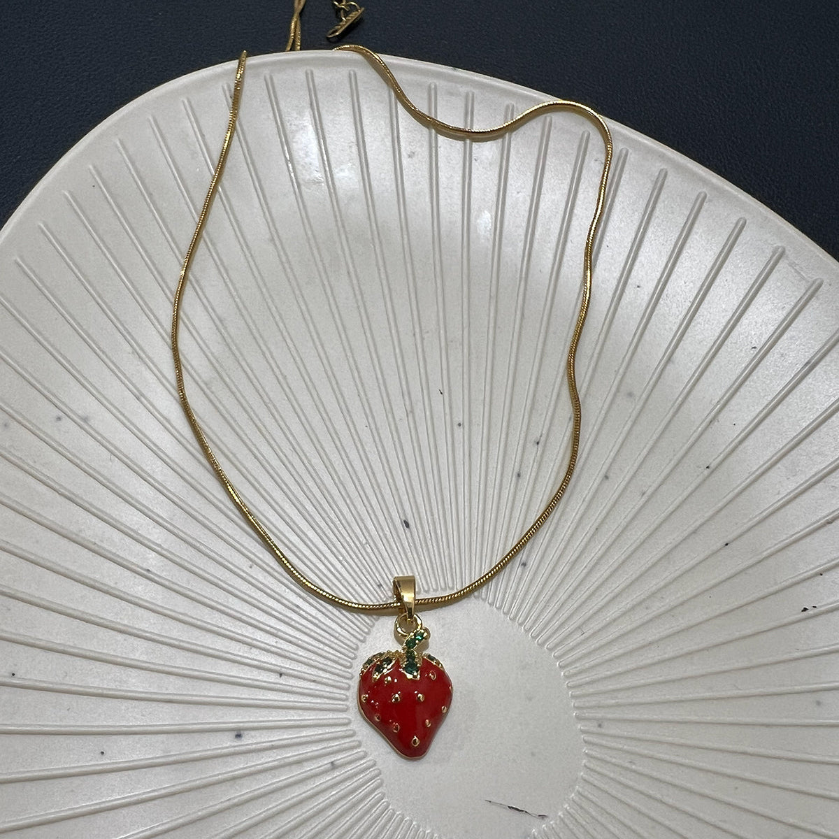 Top view of a red strawberry pendant on a gold plated snake chain styled on a white textured decorative plate.