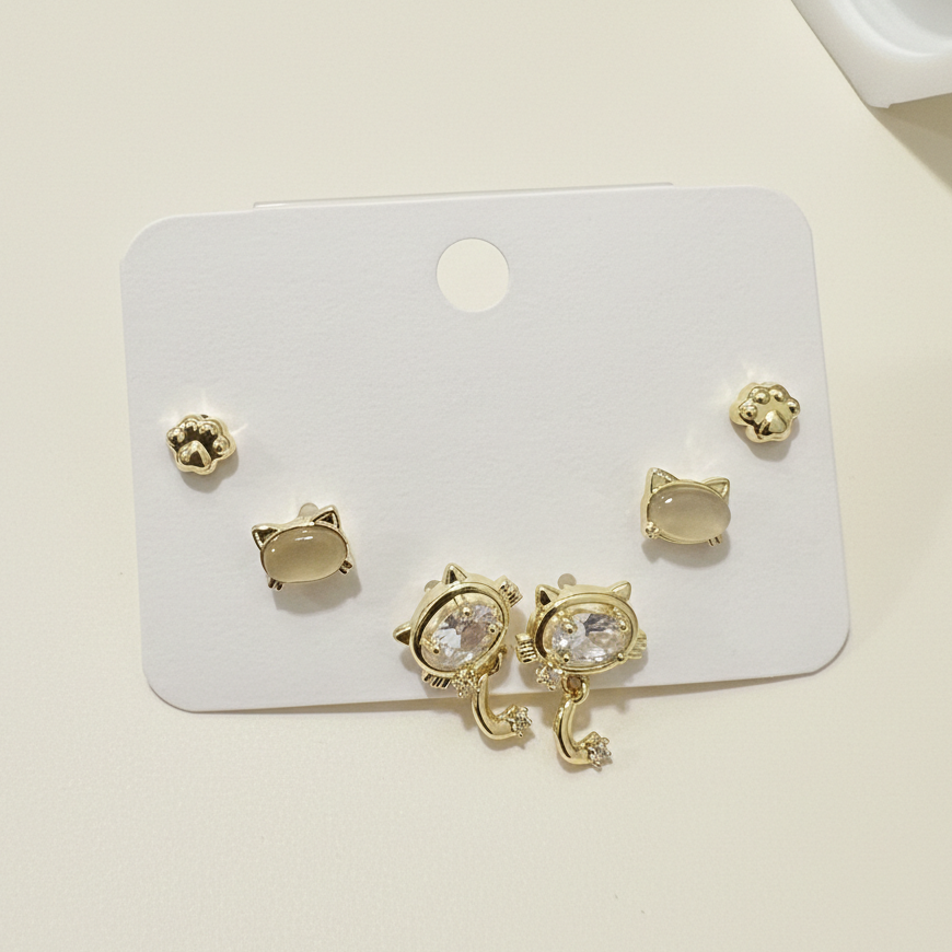 A set of three pairs of gold-plated cat-themed earrings featuring paw prints and crystal cat heads on a white display.