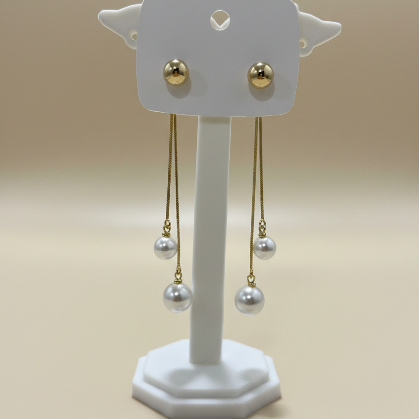 Pair of elegant pearl and gold linear earrings showcased on a white jewelry stand against a tan background