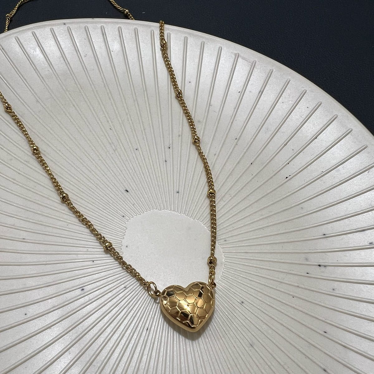 Gold plated honeycomb heart pendant necklace on a station bead chain displayed on a white textured decorative plate