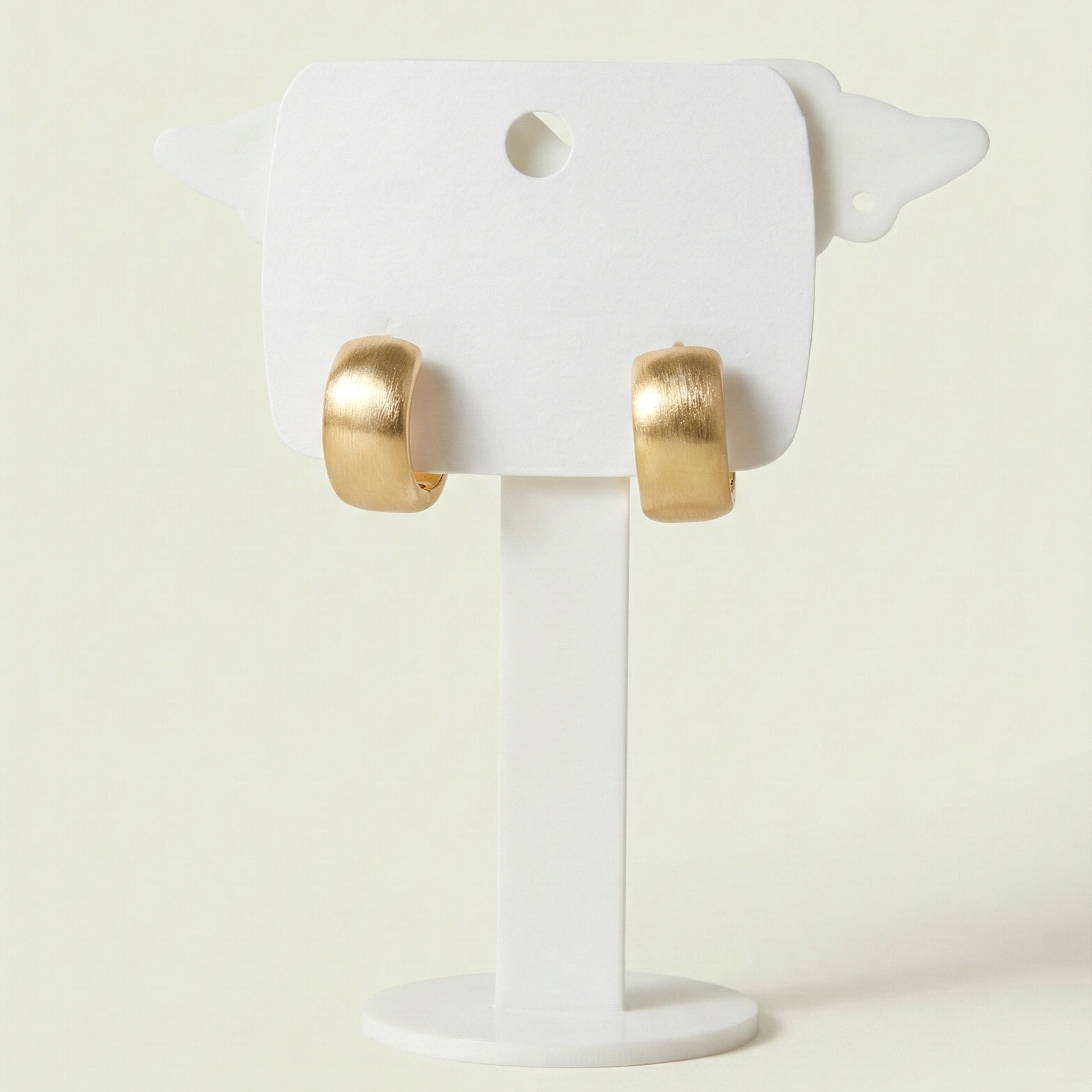 Pair of matte gold brushed hoop earrings displayed on a white stand showing the wide modern design.