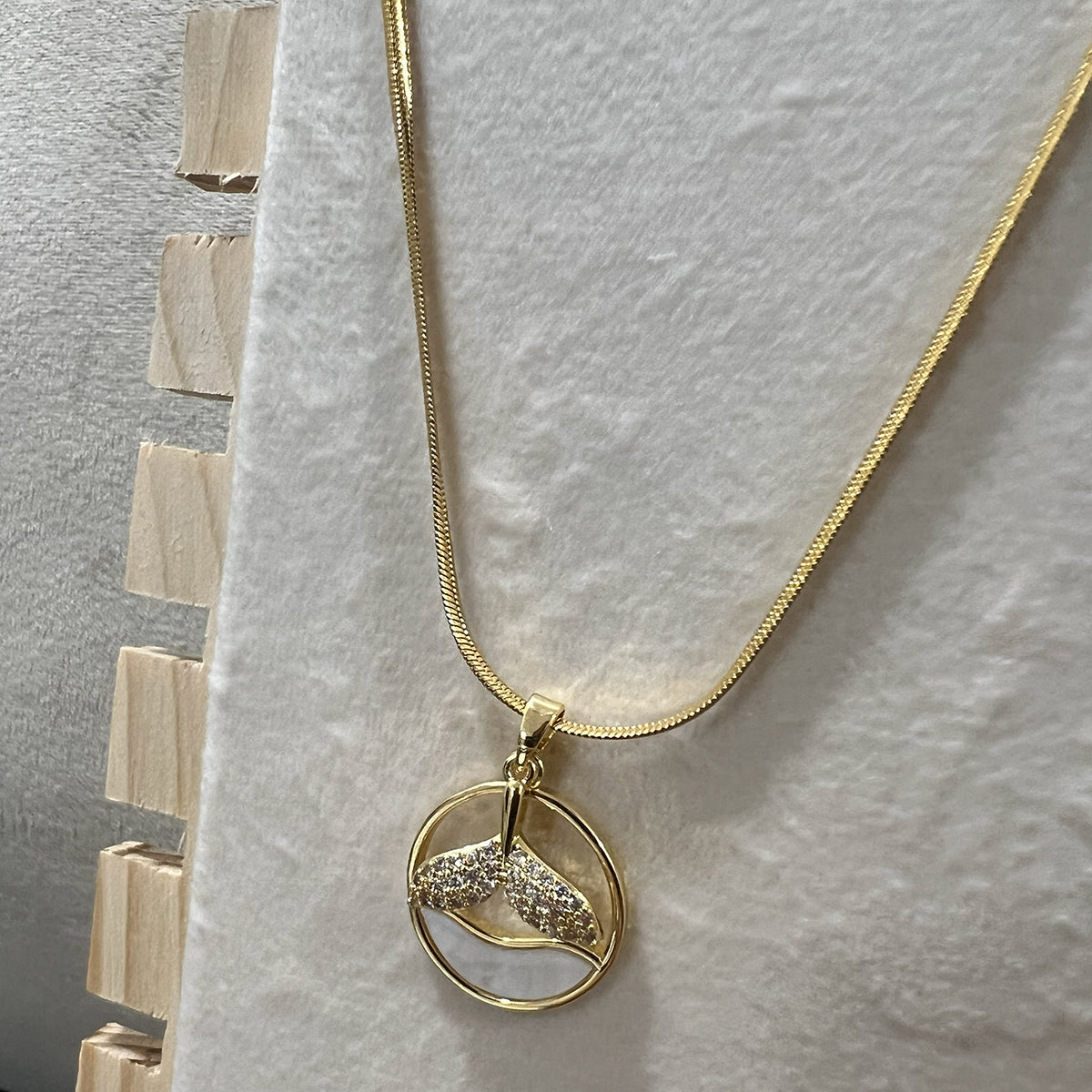 Close-up of a circular mermaid tail pendant in a gold finish displayed on a white textured stand.