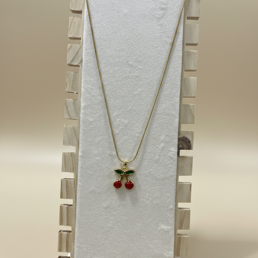 A vibrant red and green cherry pendant on a sleek gold snake chain displayed on a white textured plate