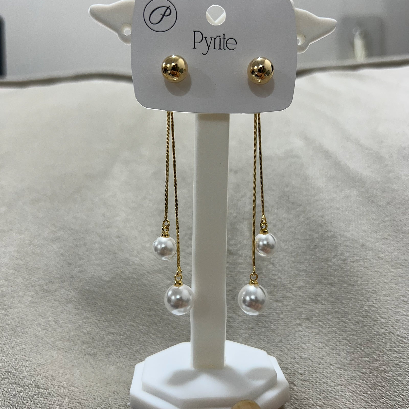 Pair of elegant pearl and gold linear earrings showcased on a white jewelry stand against a tan background
