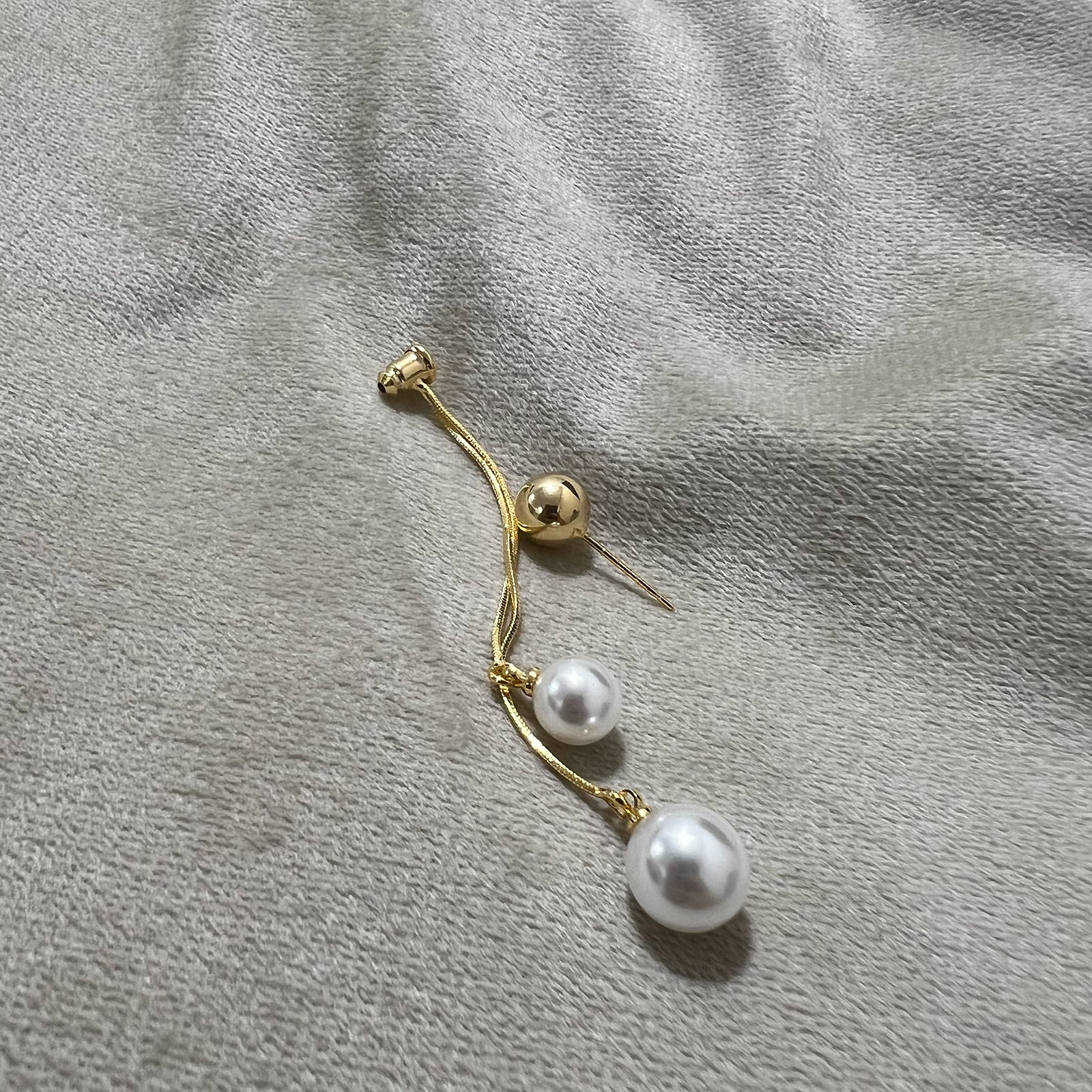 Single gold plated stainless steel drop earring with two hanging pearls on a grey textured surface
